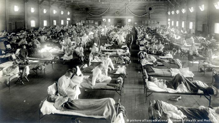 Black and white photo with a huge hall and hundreds of patients lying in beds, some accompanied by doctors wearing masks 