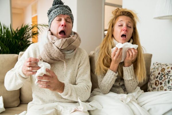 Color photography.  A couple shows a man and a woman sitting on the sofa in warm clothes and sneezing.