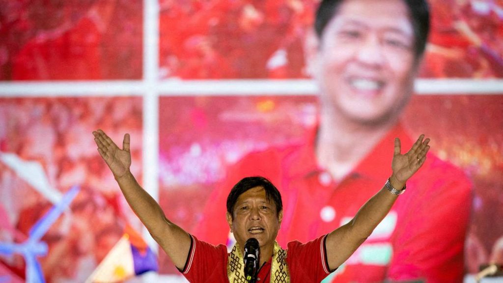 Presidentkandidat Ferdinand Marcos står med armene hevet. Han står på en scene med en mikrofon foran seg. Han har på rød t-skjorte, bak ham er det en stor skjerm, også farget rødt. Et stort bilde av ham som smiler fyller deler av skjermen.