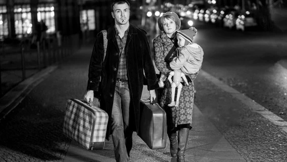 Black and white photo of a man and woman walking down the street with suitcases and a baby in their arms.  © Zeitsprung Images / Wild Bunch Germany Photo: Peter Hardwick