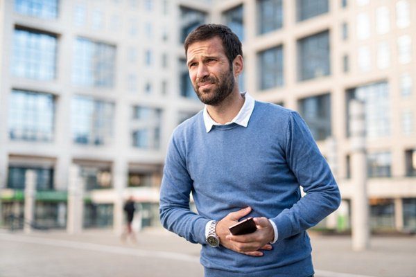 A man standing with one hand on his stomach and the other holding a mobile phone.  He wears a blue shirt, has dark hair and a beard - Metropolis