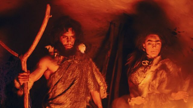 The color image shows actors with long hair and fur clothes inside a cave